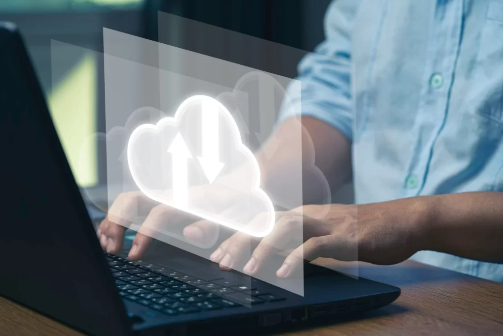 Homem de camisa azul em frente a um notebook e um símbolo digital de nuvem sobre o notebook representando um sistema em nuvem.